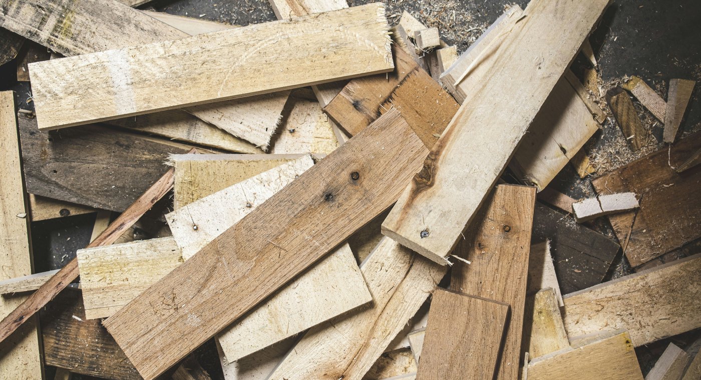 Scrap boards and pallet-wood offcuts heaped on a concrete floor, sawdust between them