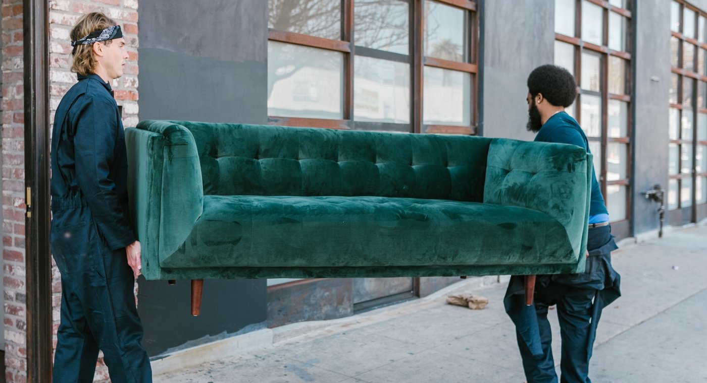 Two workers in dark coveralls carrying a green sofa out of a doorway onto the sidewalk