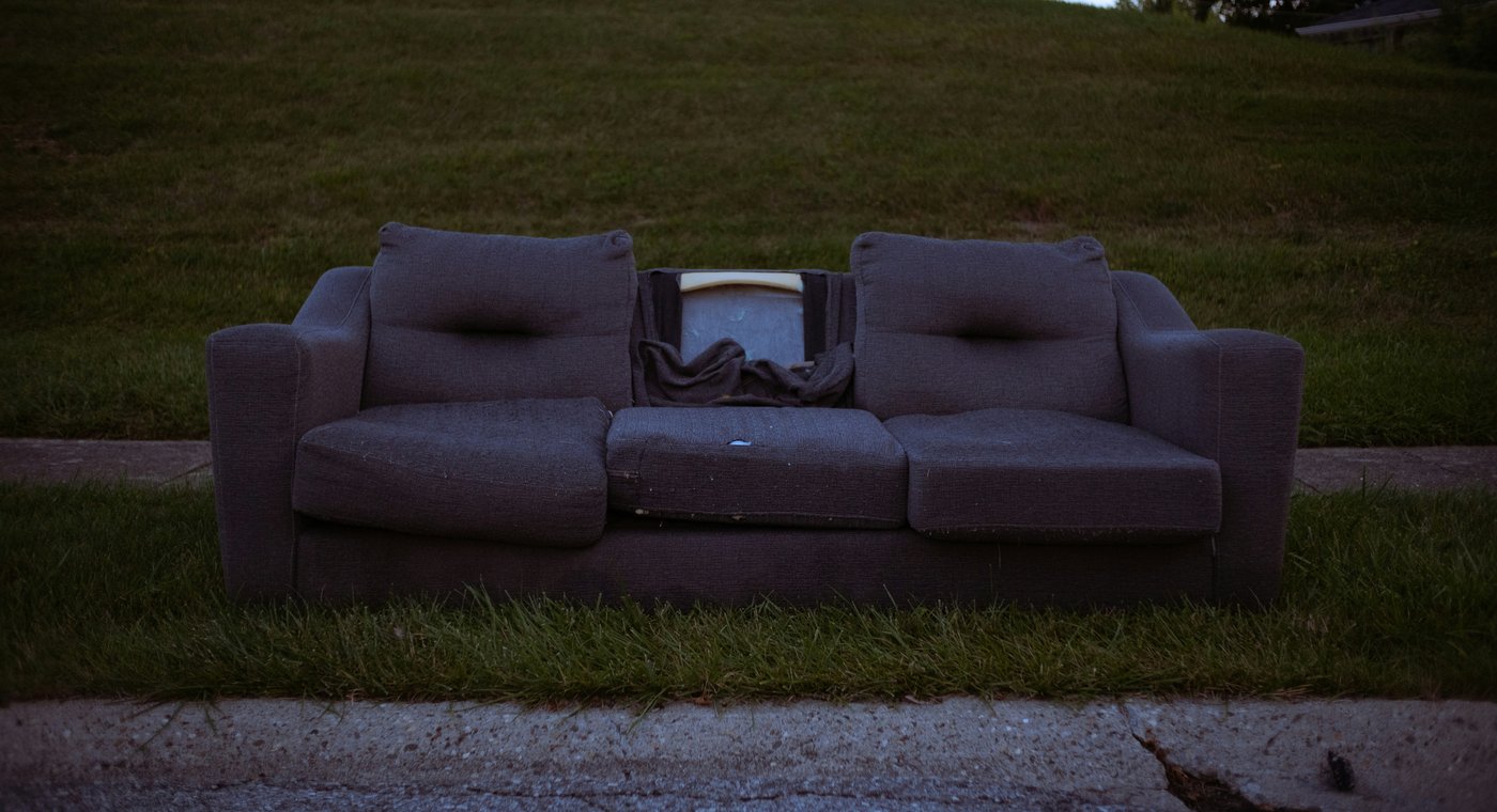 A worn gray sleeper sofa left on the grass strip between the curb and the sidewalk
