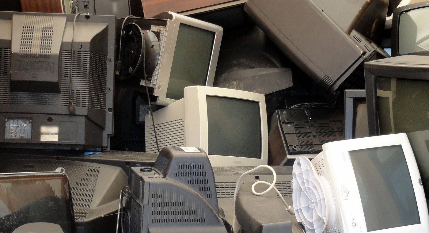 A heap of old tube televisions and computer monitors, beige and black, stacked against a plywood wall